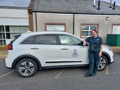 Paramedic in green uniform stood leaning on a white paramedic car that's parked in front a small building.