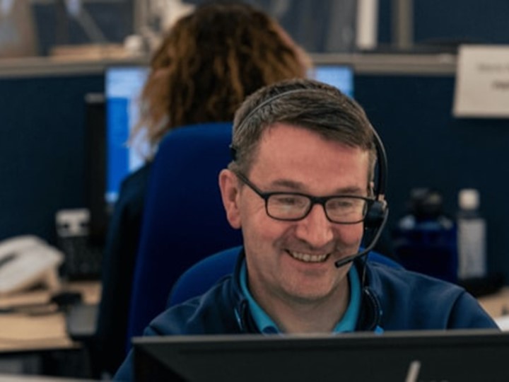 Smiling NHS 24 call handler sat at a desk with a headset on.